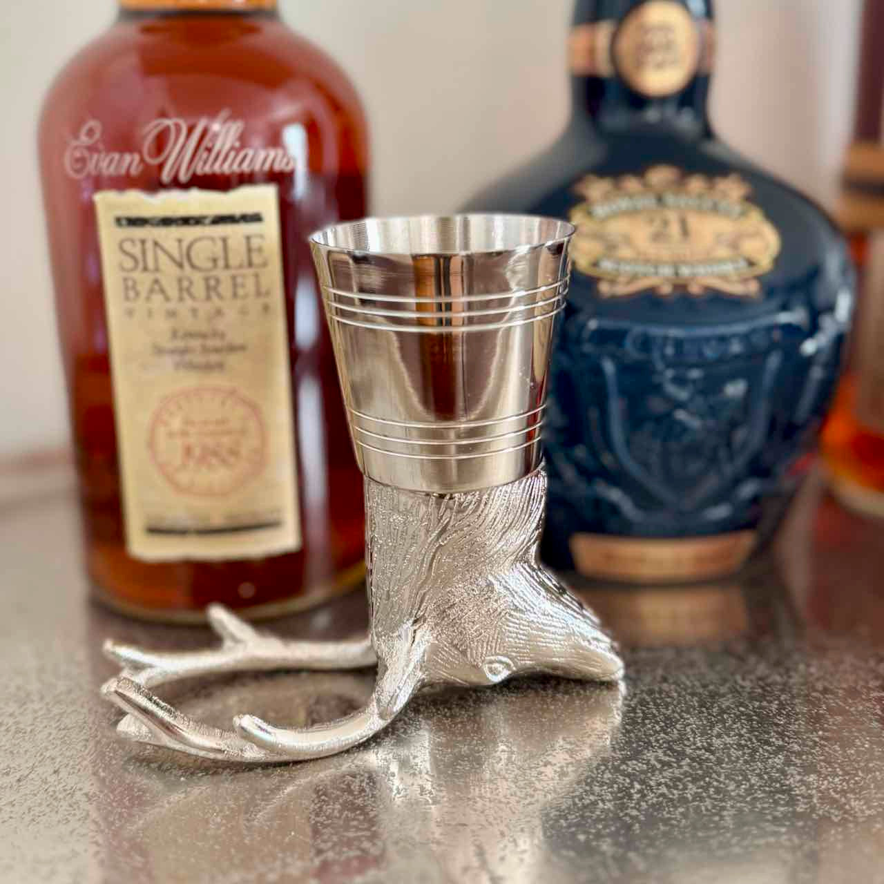 Silver deer-shaped shot glass with two bottles of whiskey in the background.