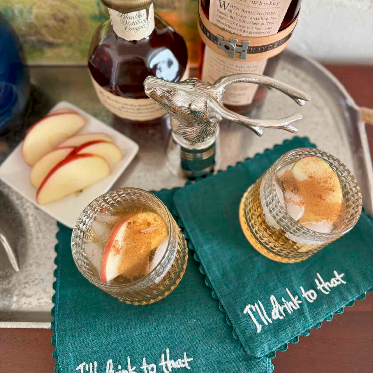 Two glasses with apple slices, a tray with bottles and snacks, and a decorative napkin with text.