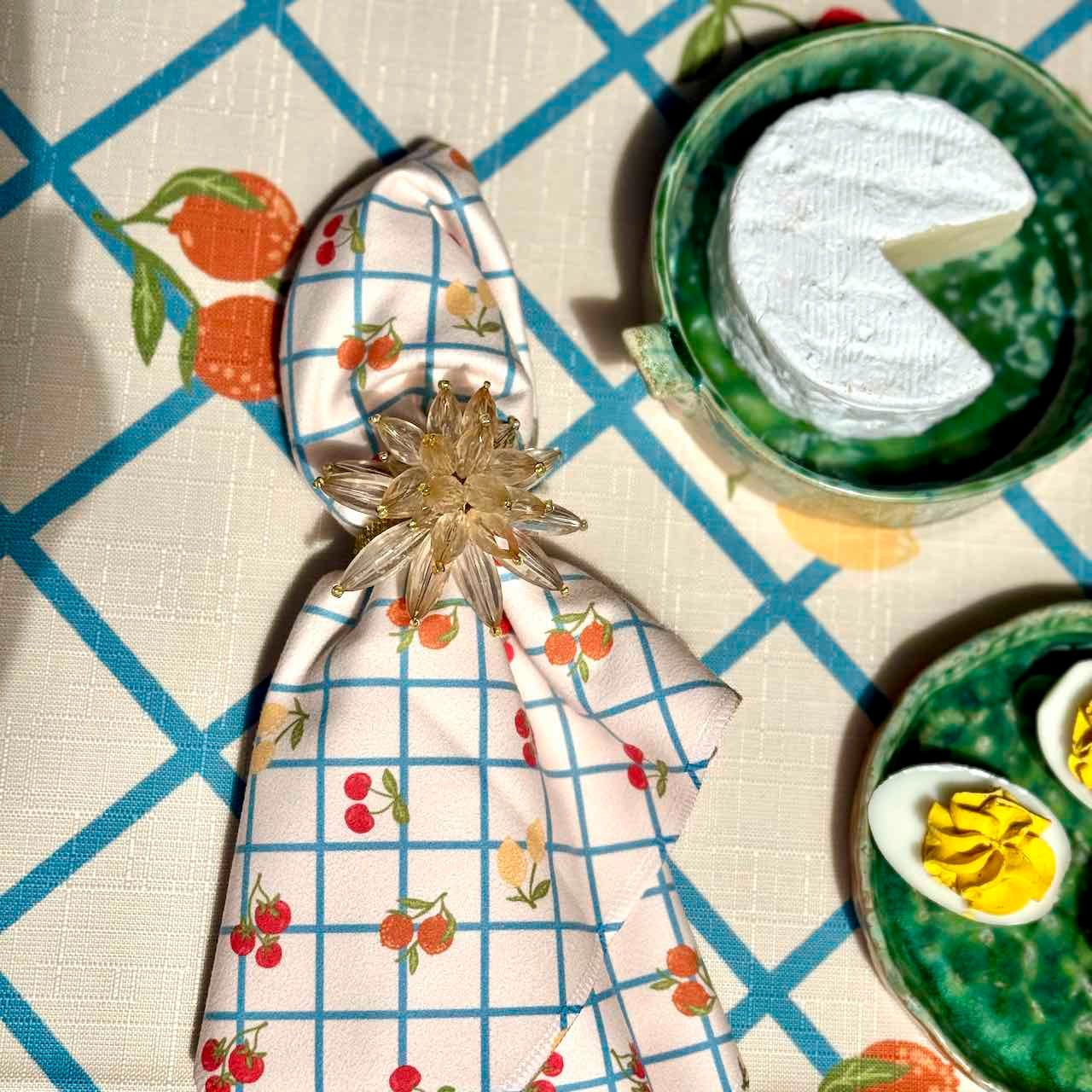 A folded patterned napkin with a cherry and grid print, secured with a clear faceted star-shaped napkin ring, placed on a table setting with green ceramic dishes and food props.