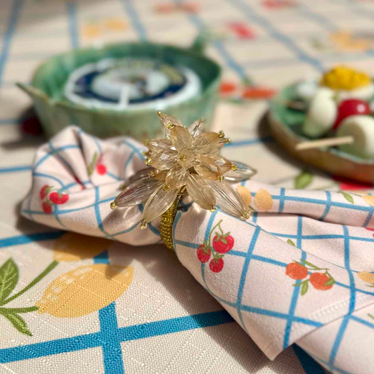 A close-up of a patterned napkin with blue grid lines and red cherries, secured with a clear crystal flower-shaped ring holder on a tablecloth with a matching fruit and floral print.