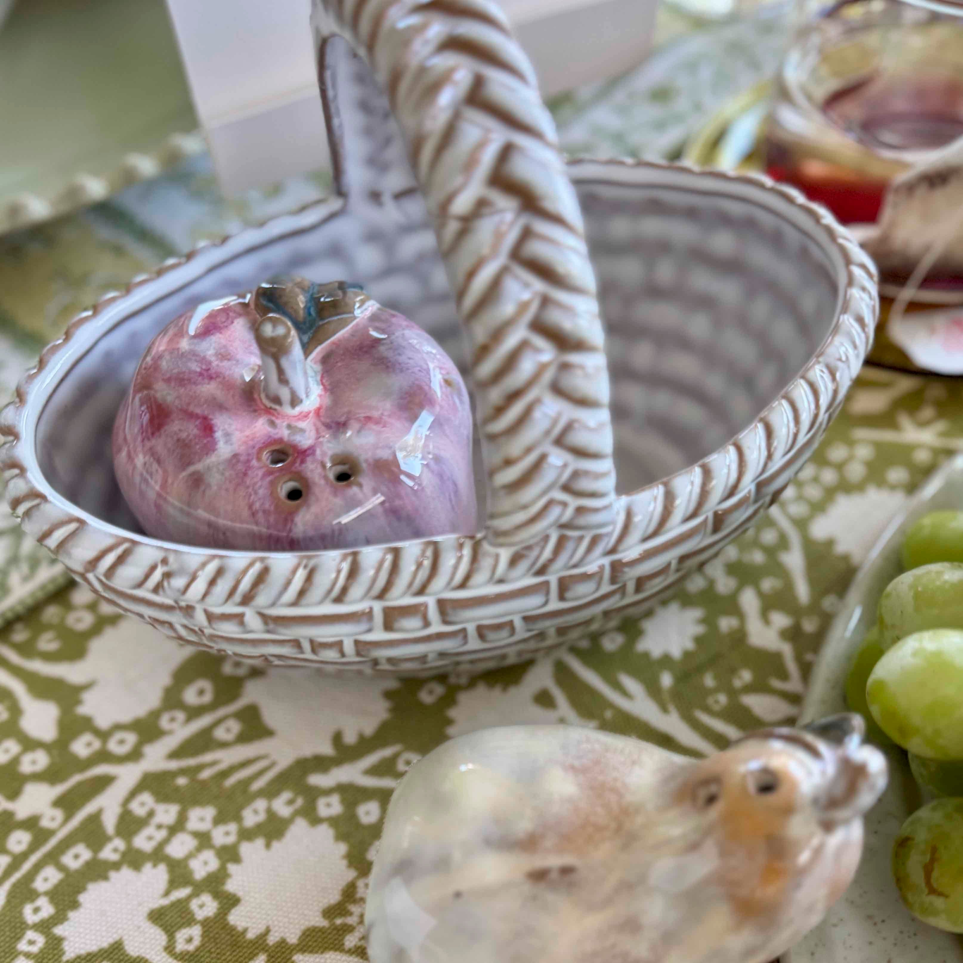 Decorative ceramic basket with a pink apple-shaped object on a patterned tablecloth.