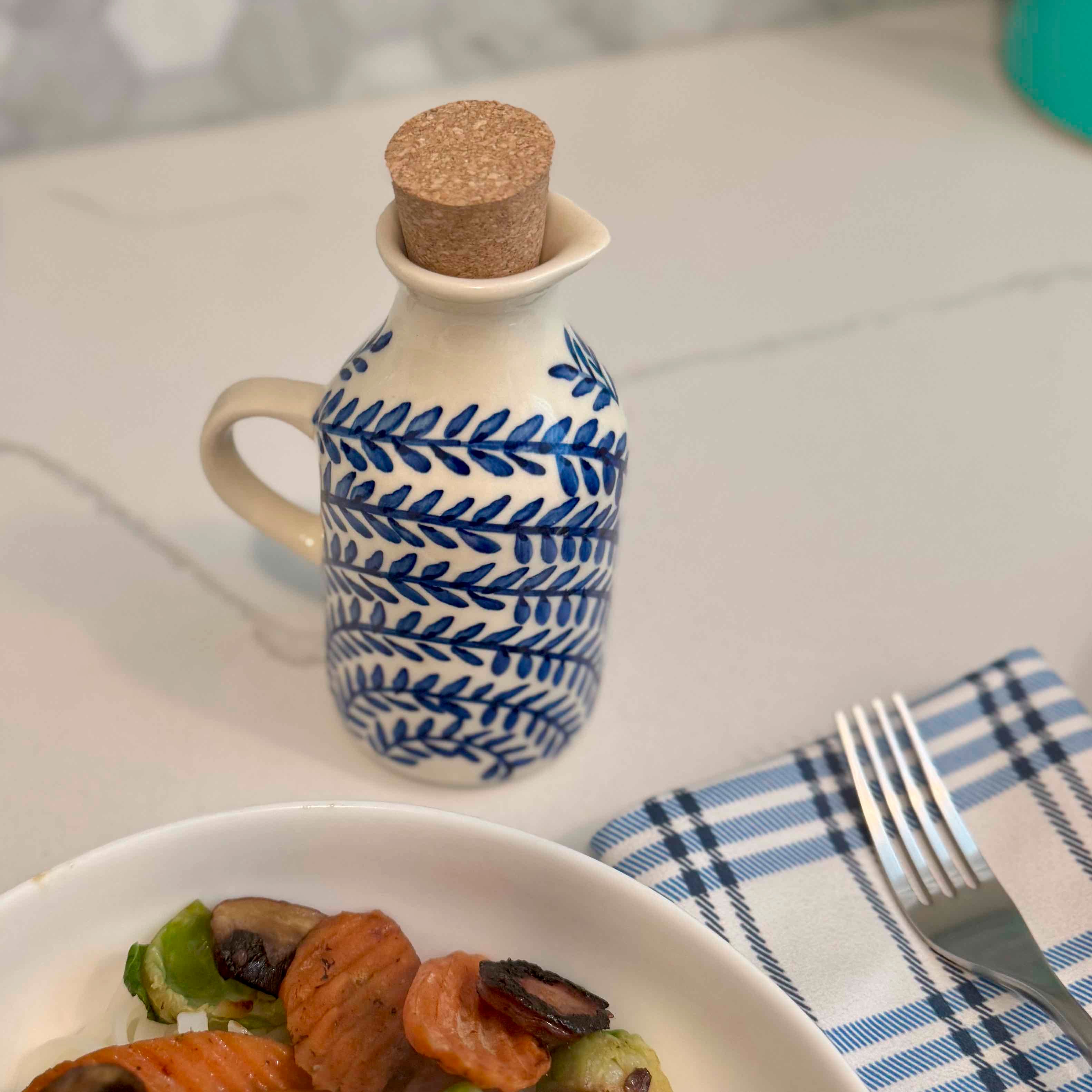 Ceramic oil bottle with blue patterns and cork lid on a table with a plate of food and fork.
