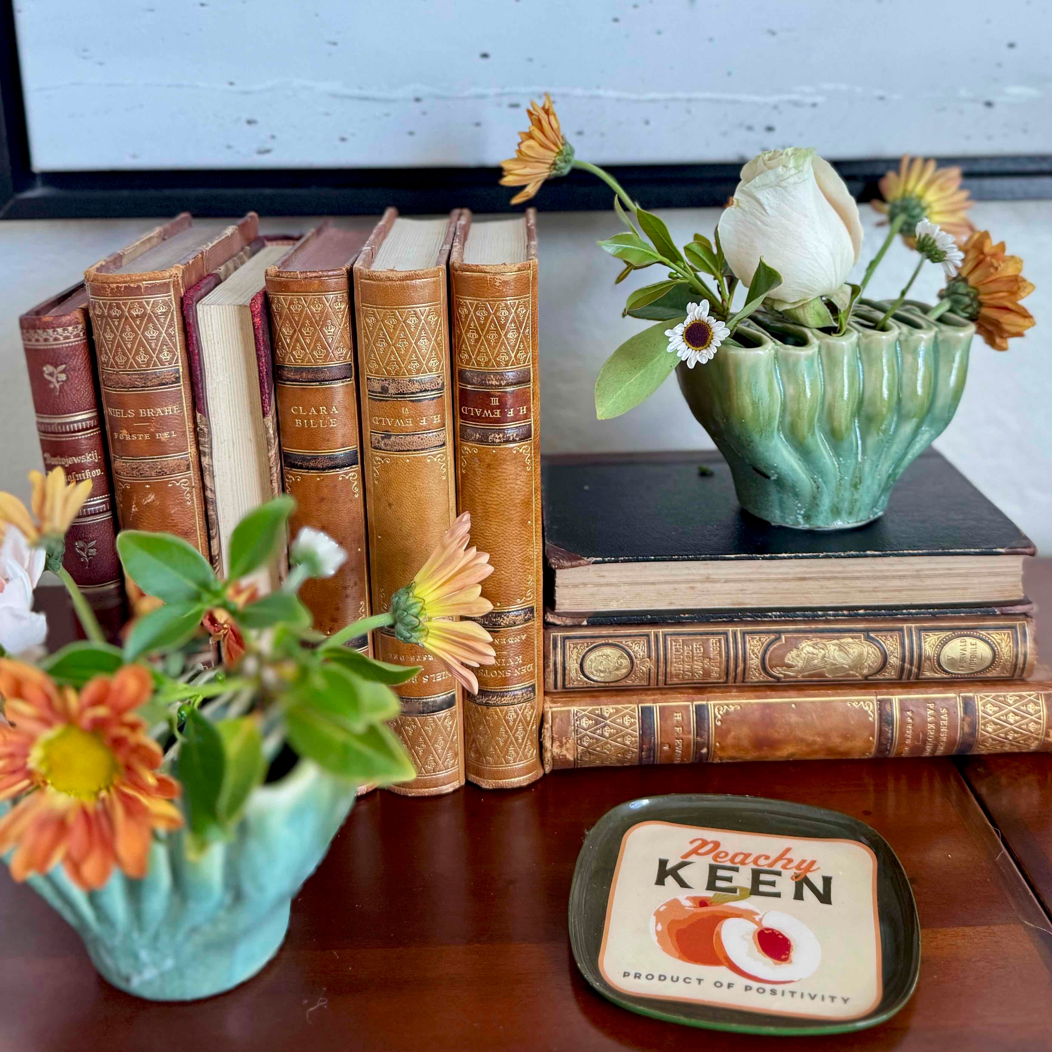 Decorative setup with vintage books, flower pots, and a 'Peachy Keen' coaster on a wooden surface.