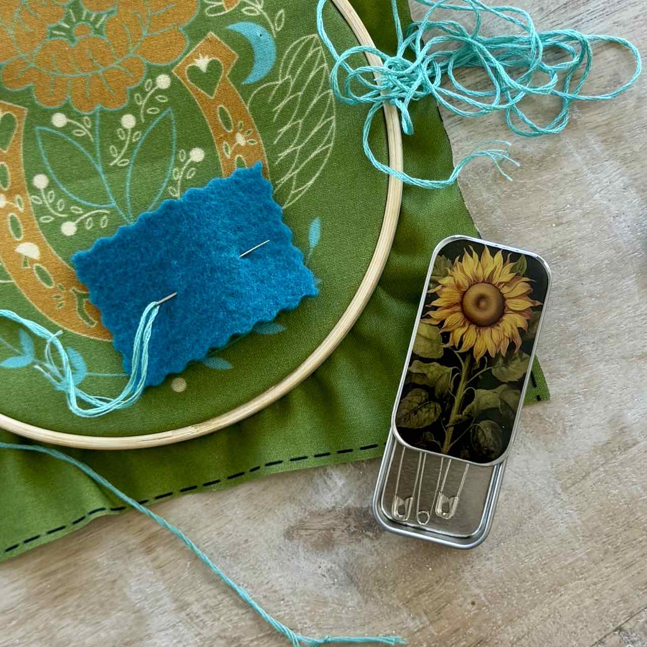 A close-up of sewing and embroidery tools on a wooden surface, including a green fabric hoop with floral embroidery, a blue felt sample, turquoise thread, and a small metal tin with a sunflower illustration and pins.