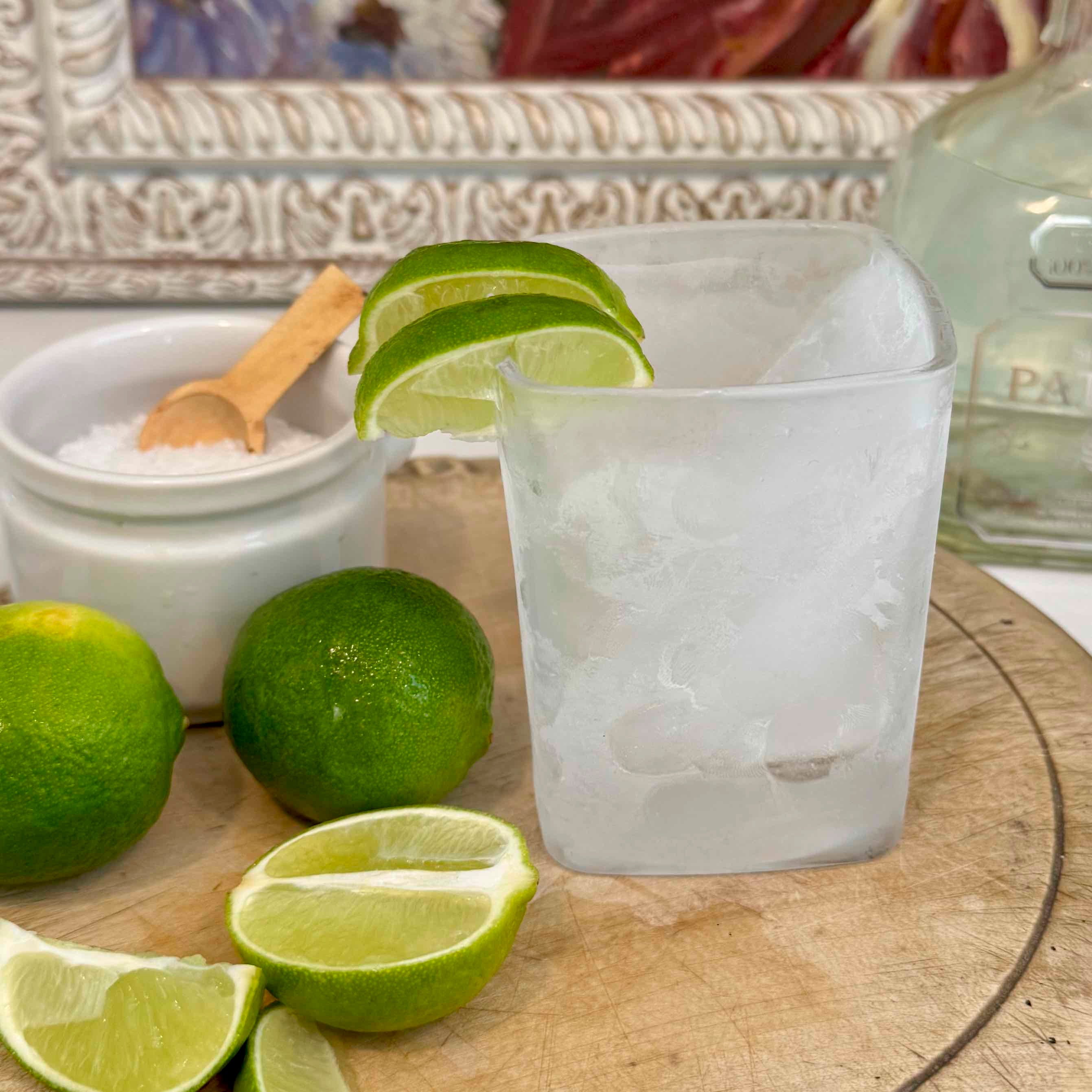 Lime wedges and a glass with ice on a wooden surface, with a bottle in the background.