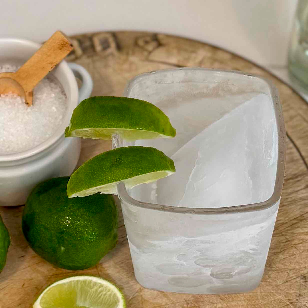 Clear glass with lime wedges on a wooden surface next to a jar of salt.