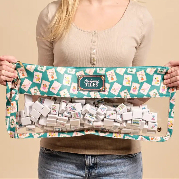 A smiling young woman holds up a clear zippered pouch filled with small white cards or blocks, featuring a teal and cream patterned fabric top with the brand name 'Madison Tiles' visible on the front label.