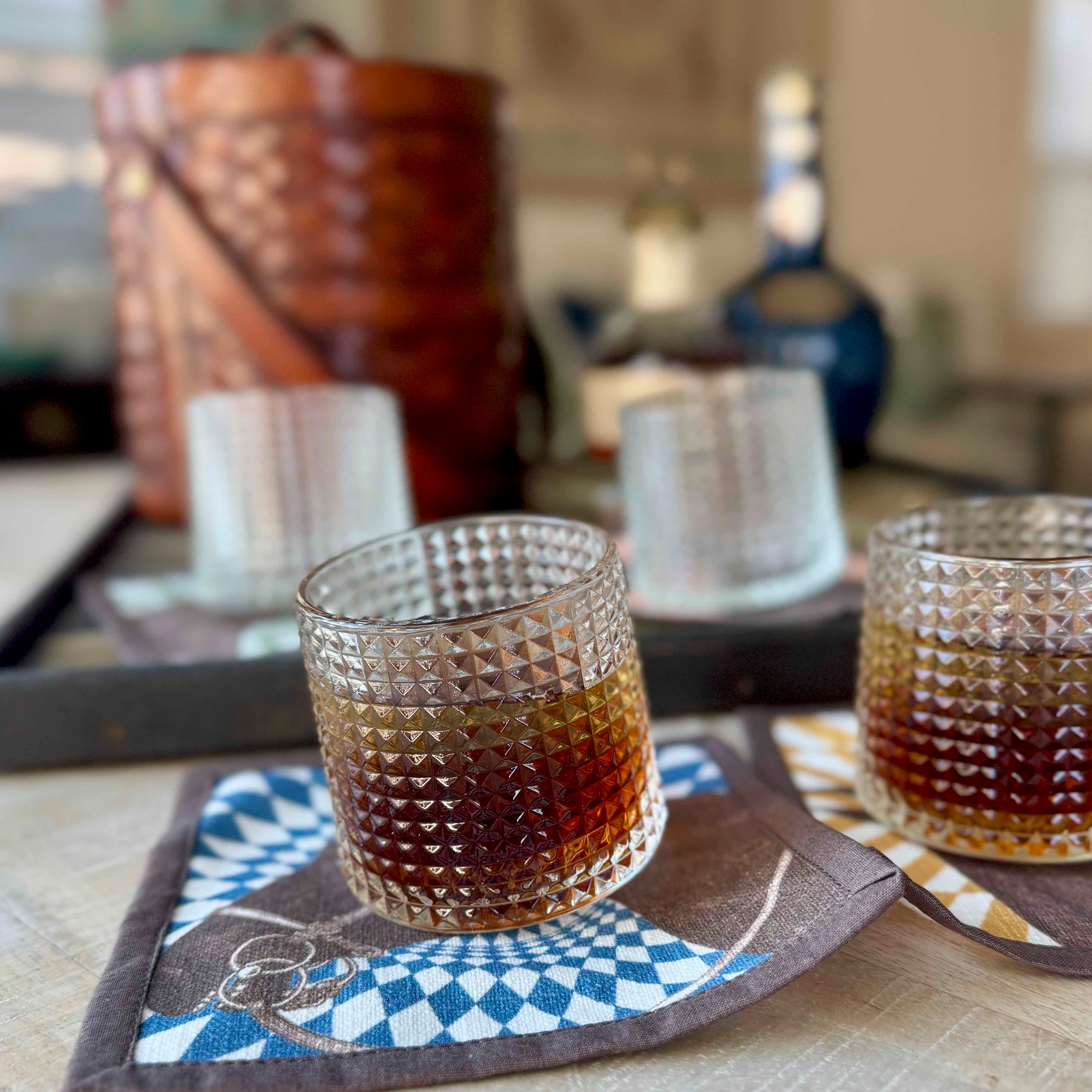 Two glasses of whiskey on checkered coasters with bottles in the background