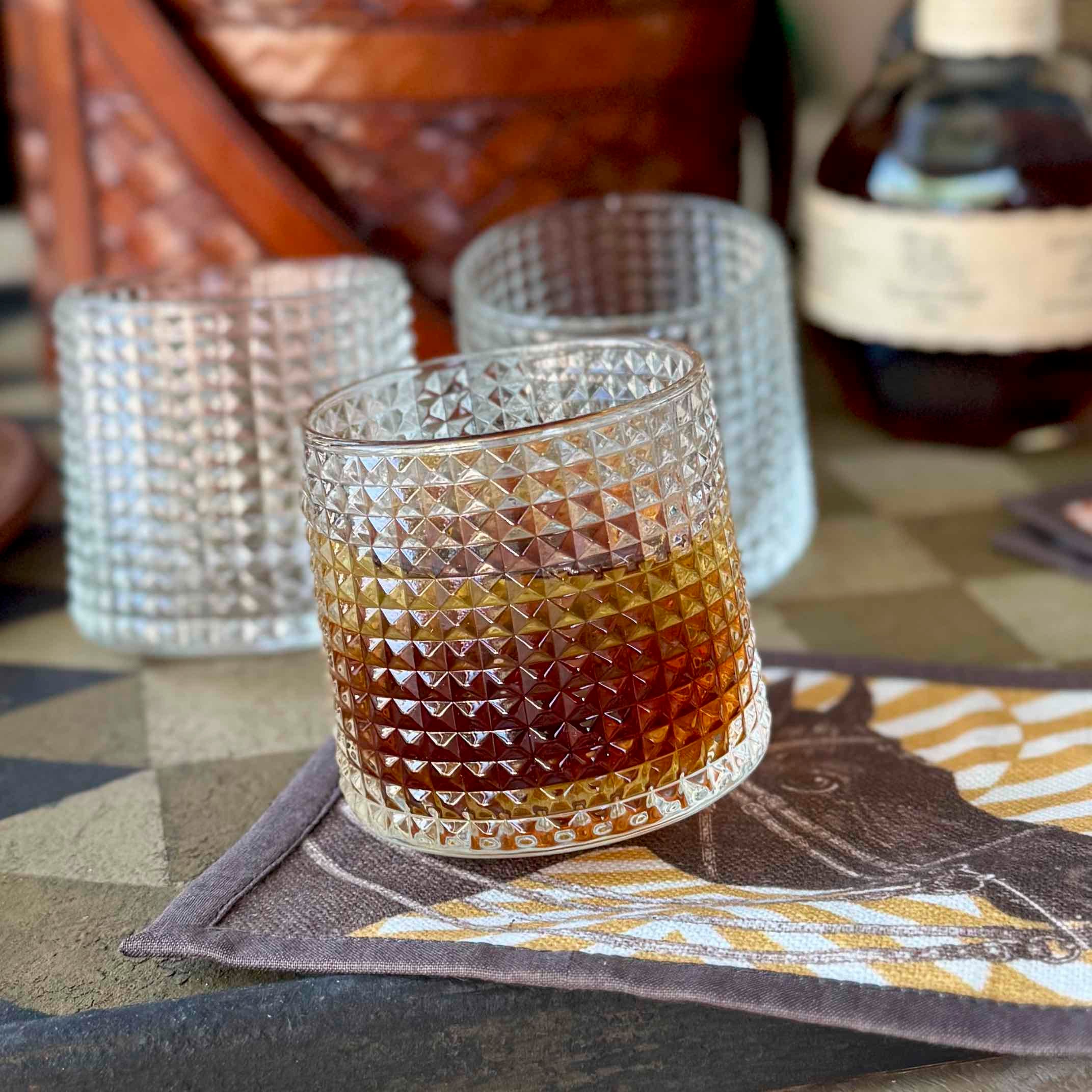 Glass with a diamond pattern filled with a layered drink on a decorative mat, with a bottle in the background.