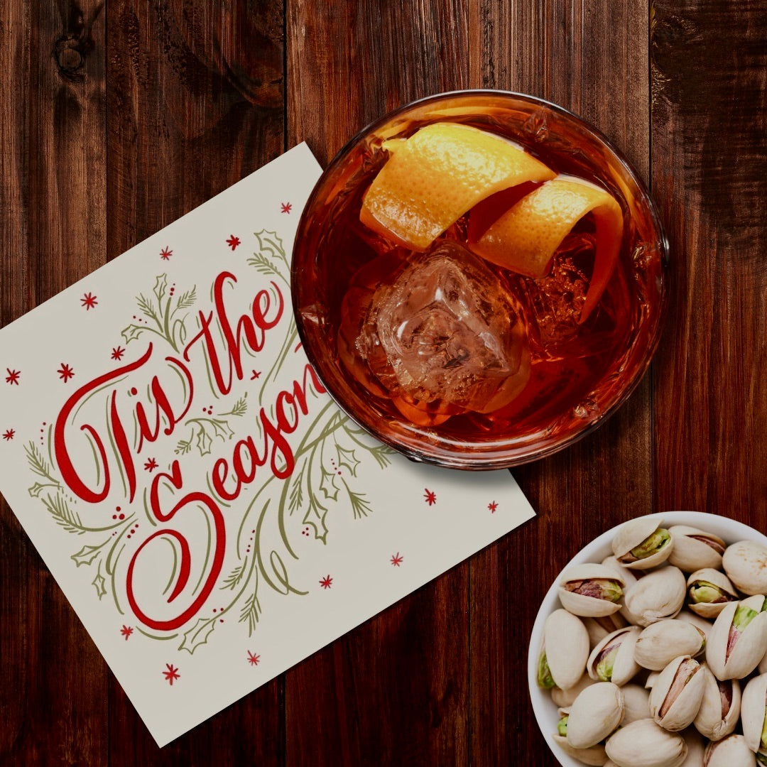 Glass of cocktail with orange peel, 'Tis the Season' card, and bowl of pistachios on a wooden surface