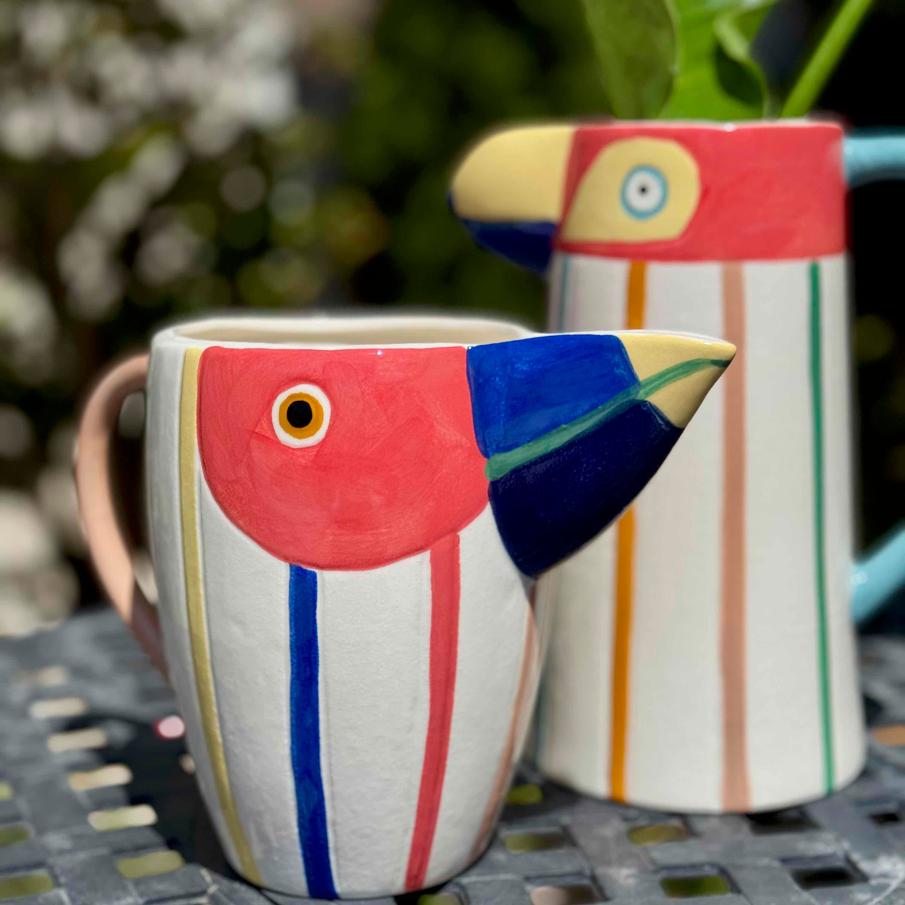 Two colorful ceramic bird-shaped mugs on a metal surface with a blurred natural background.
