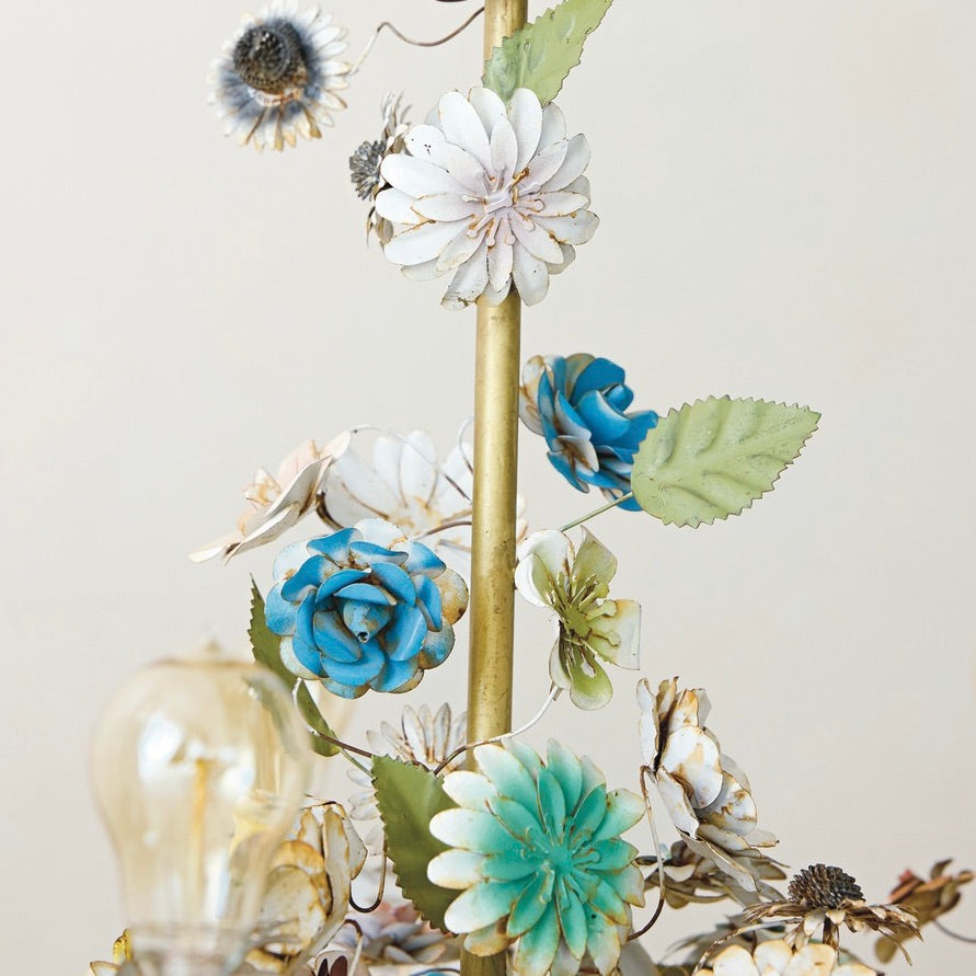 Decorative floral arrangement with blue, green, and white flowers on a gold stand against a light background.