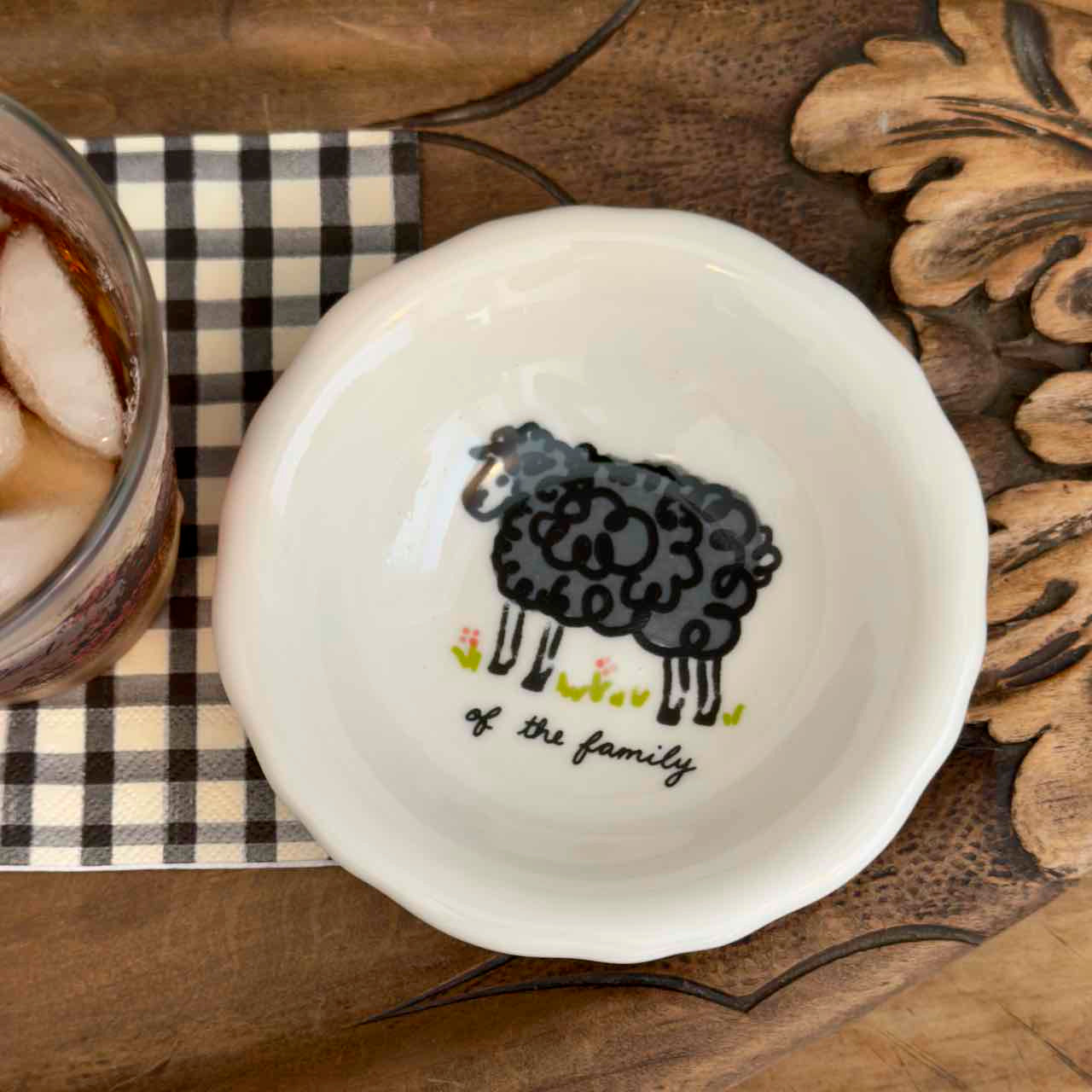 A white ceramic bowl with a scalloped rim, featuring a black cartoon sheep illustration and the text 'of the family' in black script. The bowl is styled on a rustic wooden surface next to a checkered napkin and a glass of iced tea.