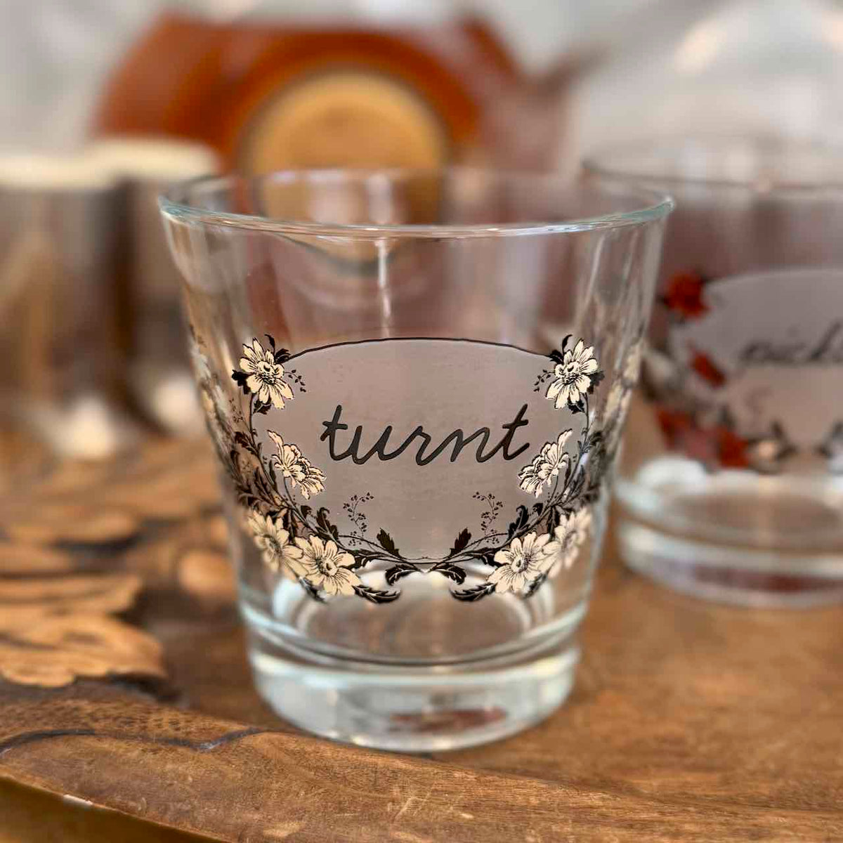 A clear glass shot glass with a frosted center label that reads 'turnt' in black script, surrounded by white and black floral illustrations. The glass is placed on a wooden surface with a softly blurred background showing other decorative items.
