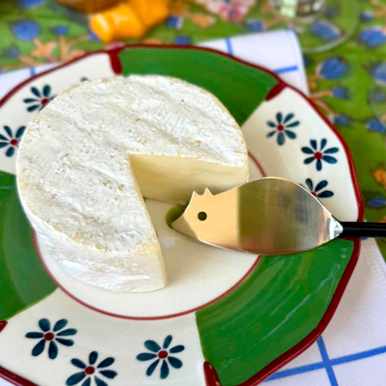 A round wheel of soft white cheese, with one slice removed, resting on a decorative green and white plate with blue flowers. A small metal cheese server with a black handle is placed on the slice.