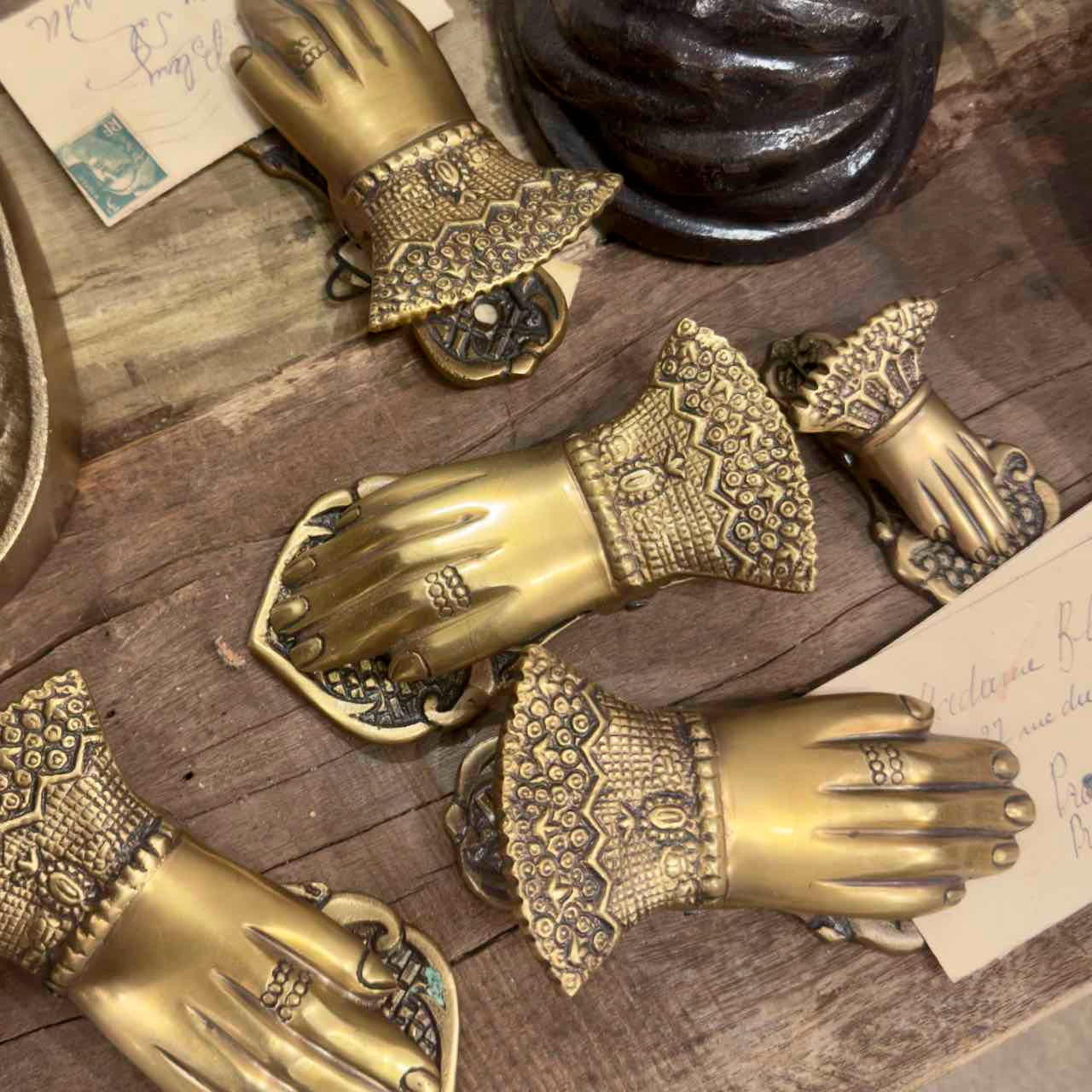 A set of ornate antique-style brass hand-shaped drawer pulls with intricate filigree cuffs, arranged on a rustic wooden surface alongside vintage papers.
