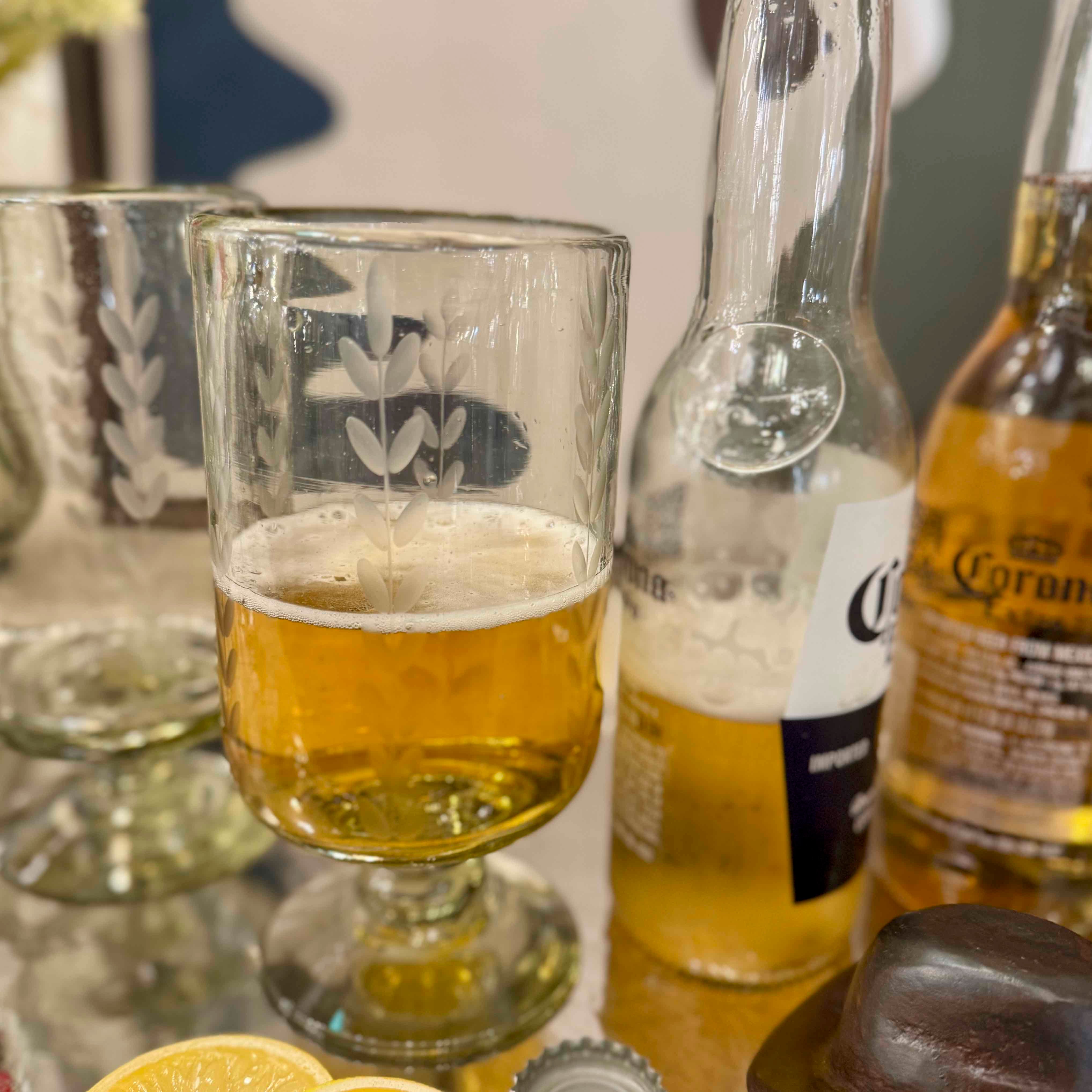Close-up of a glass of beer with Corona bottles and a lemon wedge on a table.