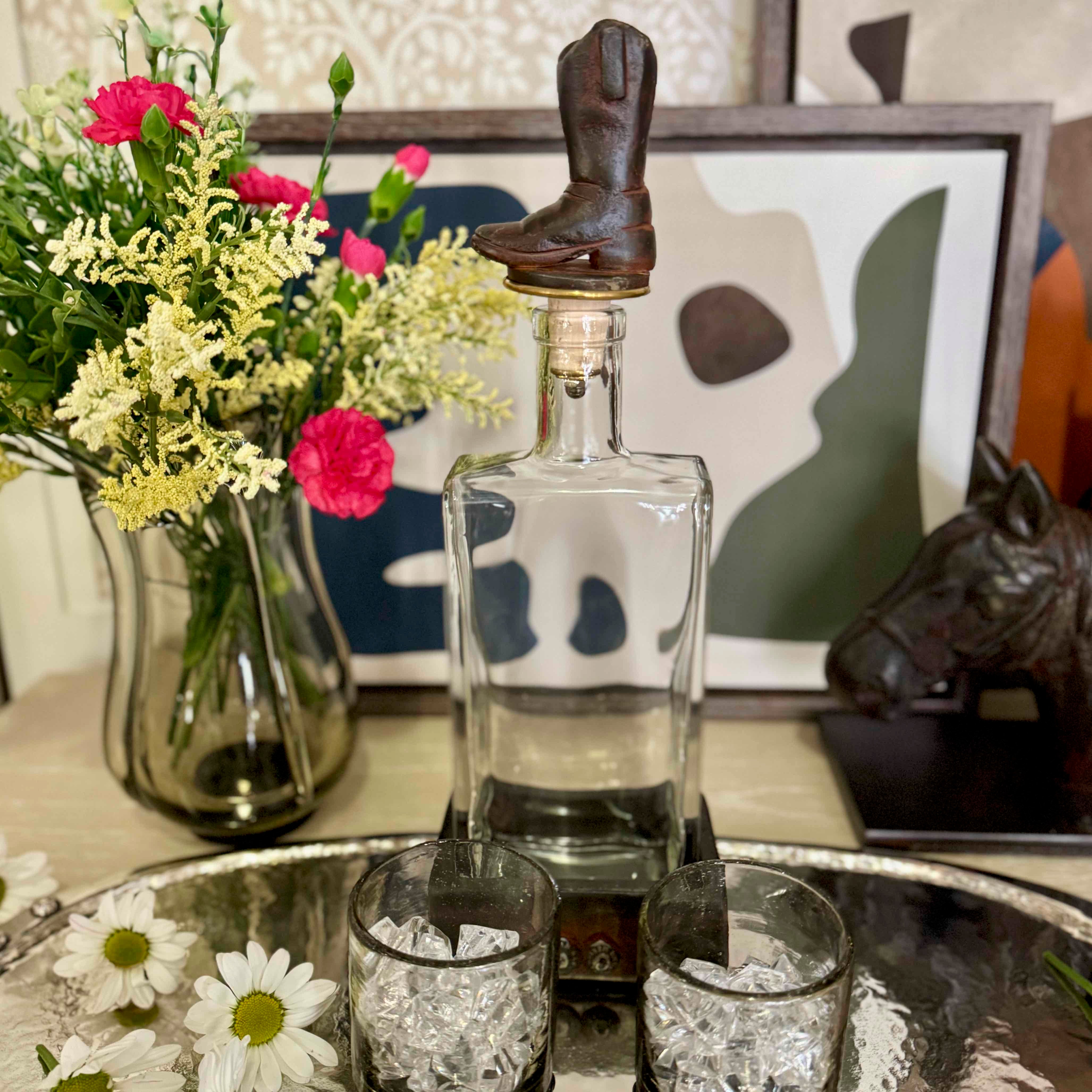 Decorative setting with a glass decanter, cowboy boot, and flowers on a table.