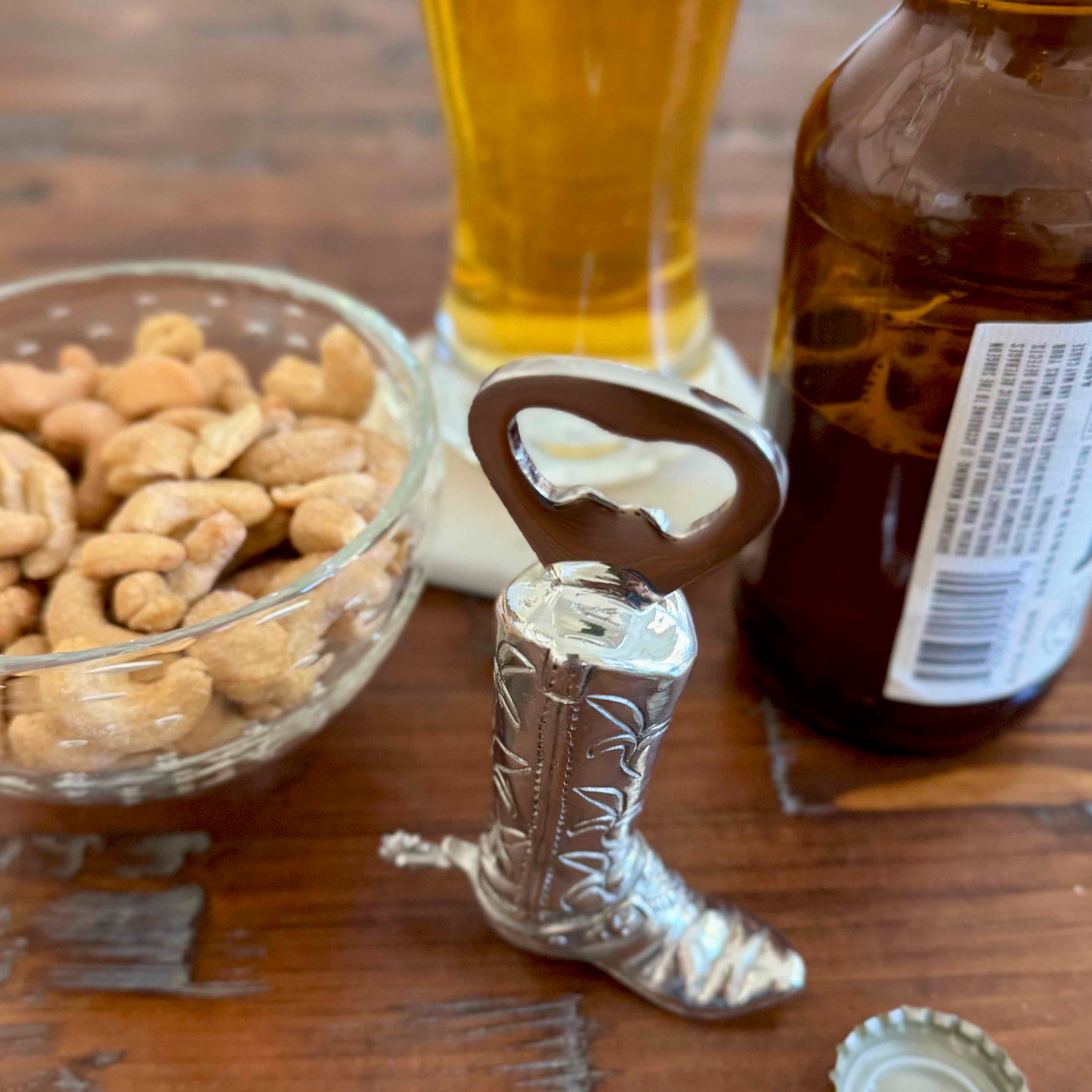 Cowboy boot bottle opener with a glass of beer, bowl of nuts, and bottle in the background on a wooden surface.