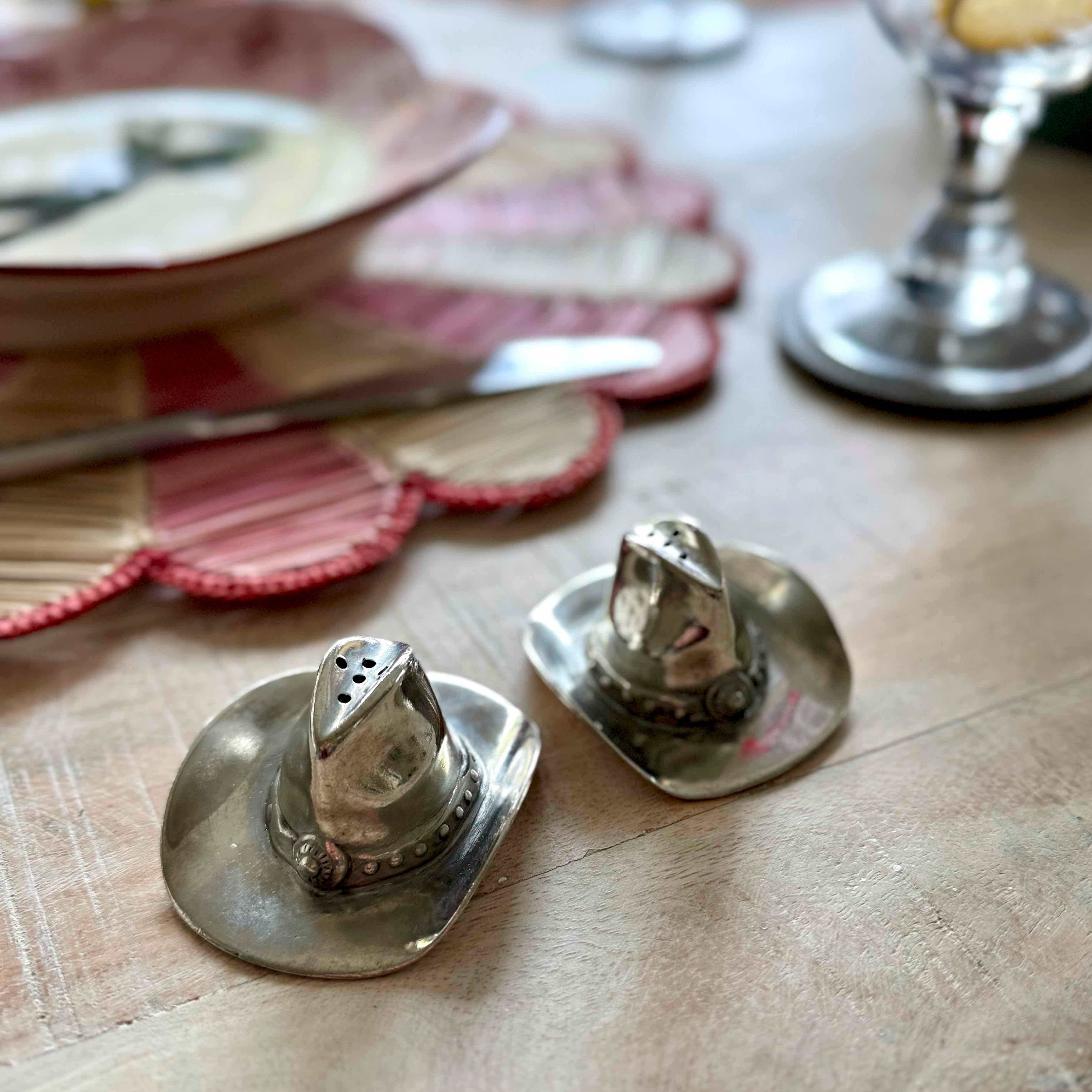 Two silver cowboy hat-shaped salt and pepper shakers on a wooden surface with books and plates in the background.