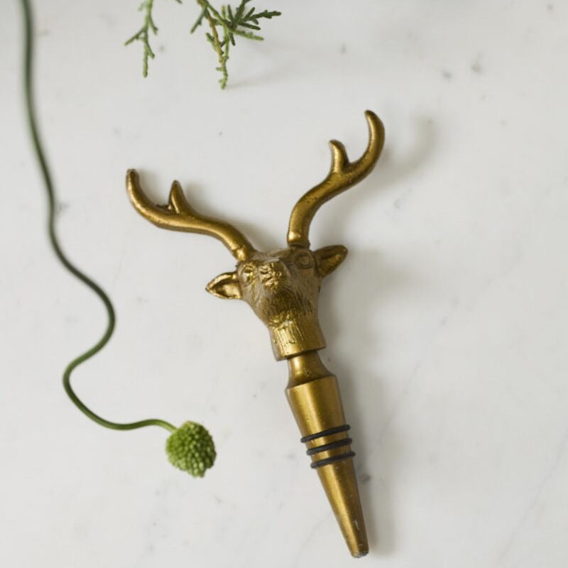 A brass deer head bottle stopper with antlers and a pointed pourer base, styled on a white marble surface with greenery and a bottle in the background.