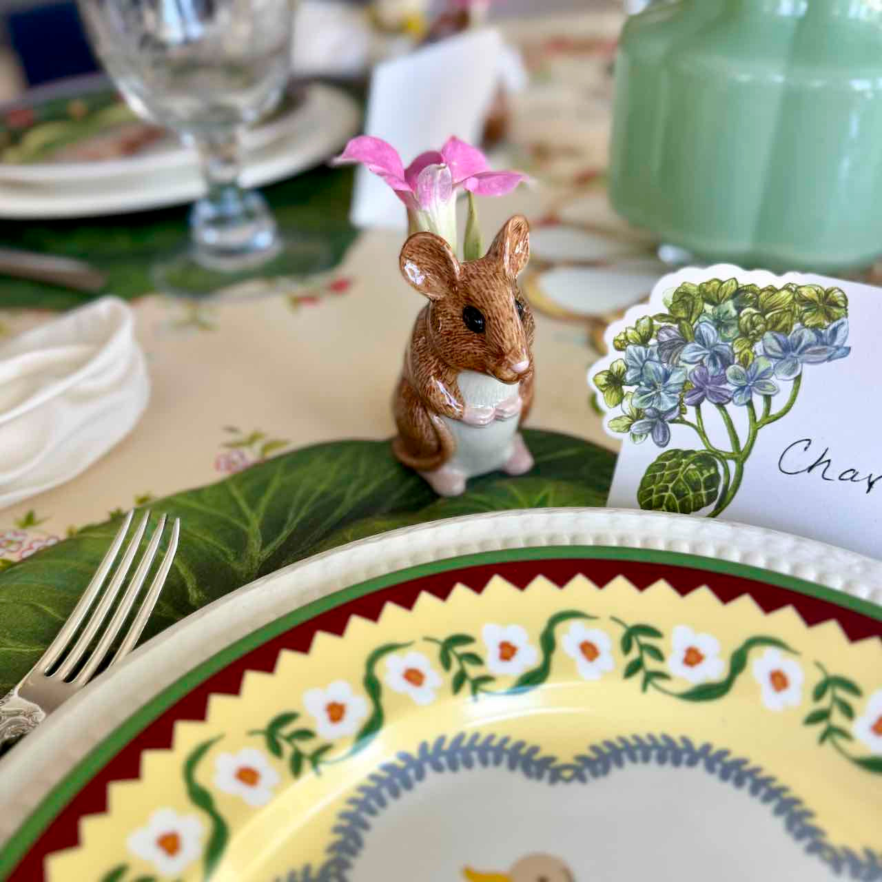 Decorative table setting with a mouse figurine, floral card, and patterned plates.