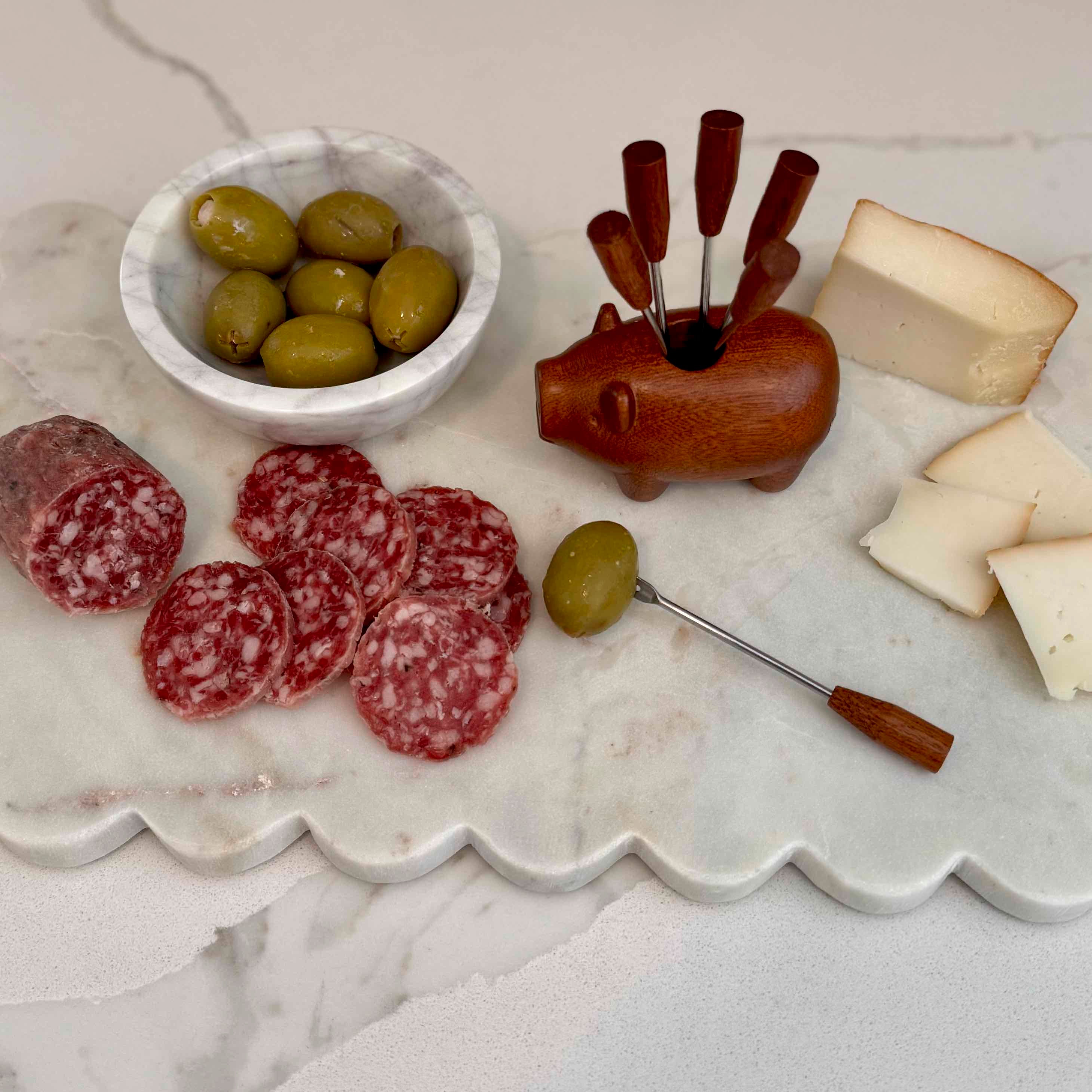 Assorted meats, olives, and cheese on a marble surface with a wooden pig-shaped appetizer set.