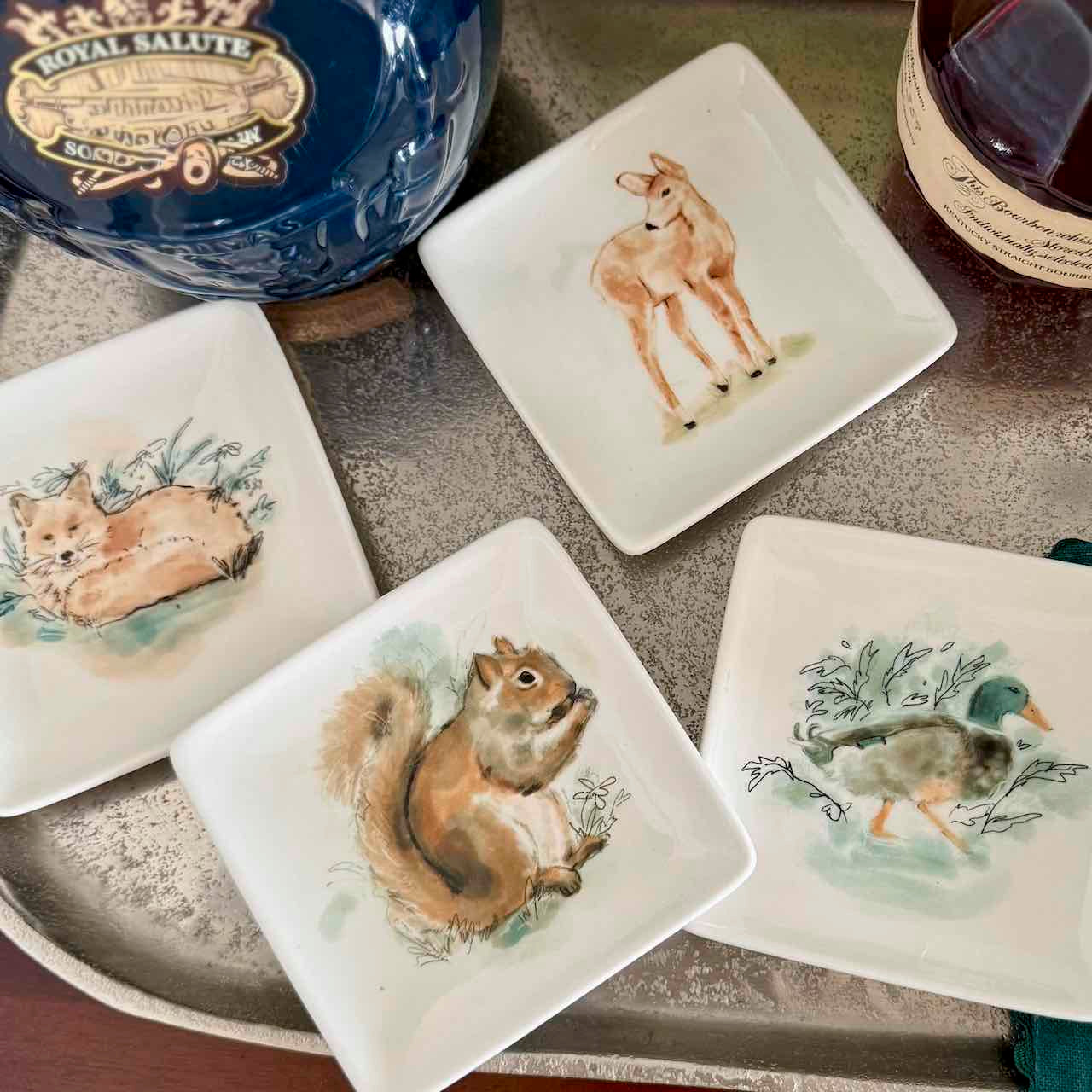 Set of ceramic plates with animal illustrations on a table with a bottle of whisky in the background.