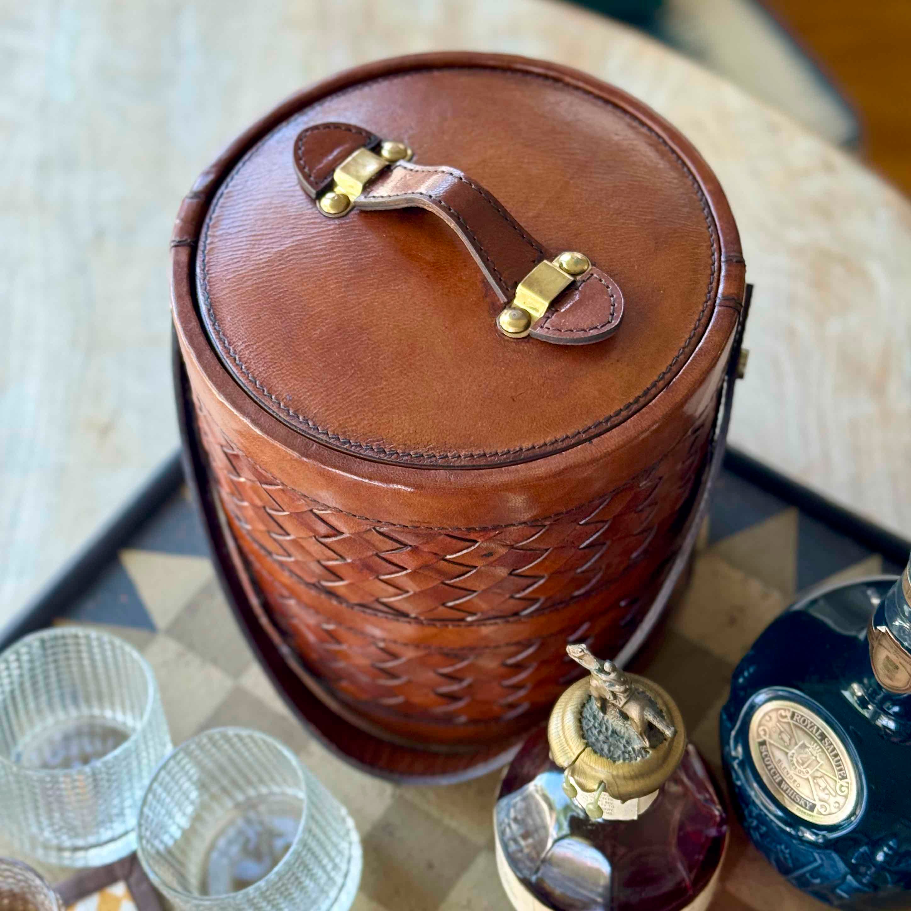 Brown leather ice bucket with woven pattern on a table with glasses and bottles.