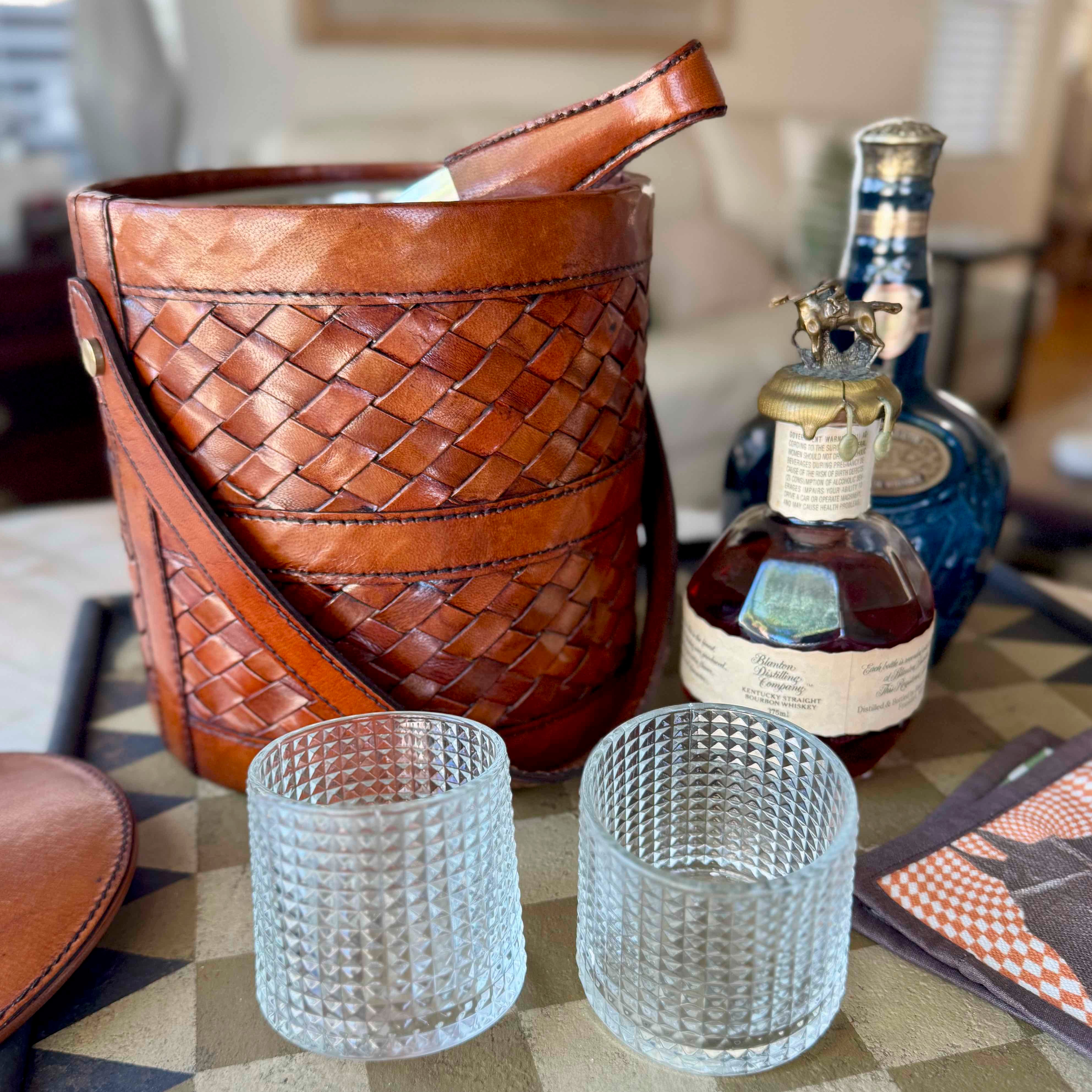 Brown woven leather ice bucket with two clear glass tumblers and a bottle of liquor on a checkered tablecloth.