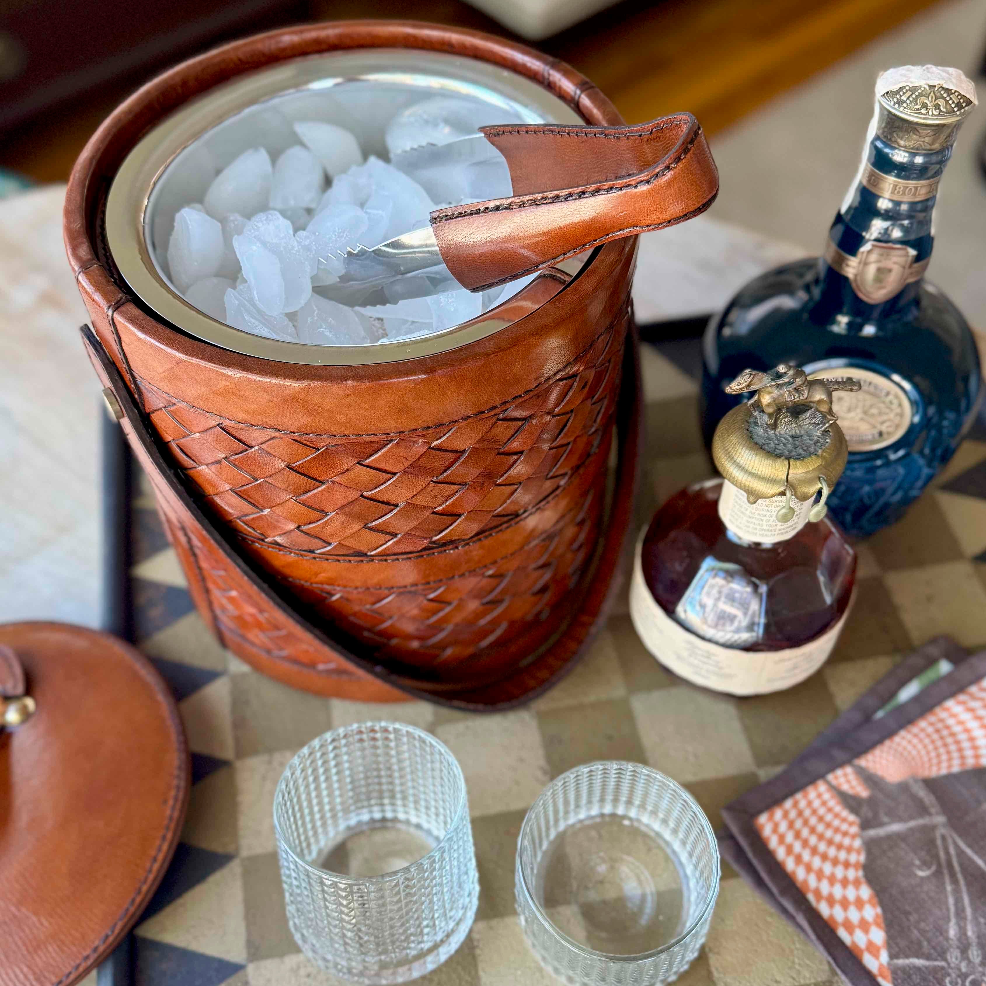 Brown woven ice bucket with ice, glassware, and bottles on a checkered tablecloth.