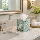 A decorative tissue box cover filled with white tissues sits on a marble bathroom counter beside a soap dispenser, mirror, and flowers.