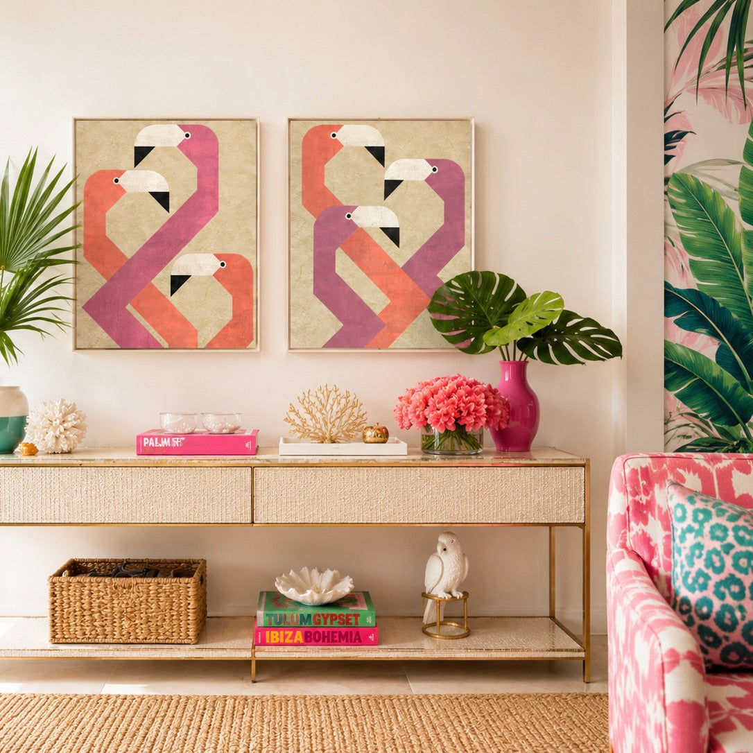 A styled interior scene showing two framed abstract flamingo wall art prints above a console table, with plants, books, coral decor, and a pink patterned chair in a real room setting.