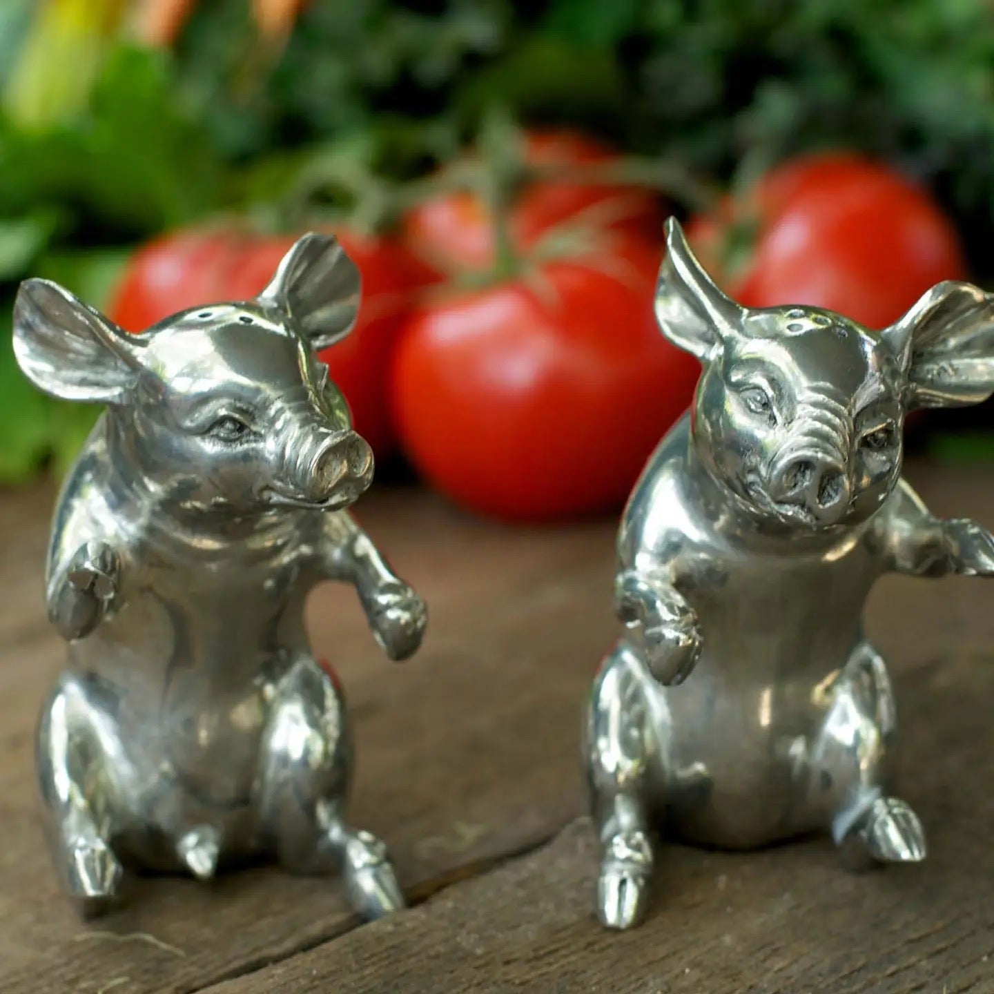 Two silver pig figurines on a wooden surface with tomatoes in the background.