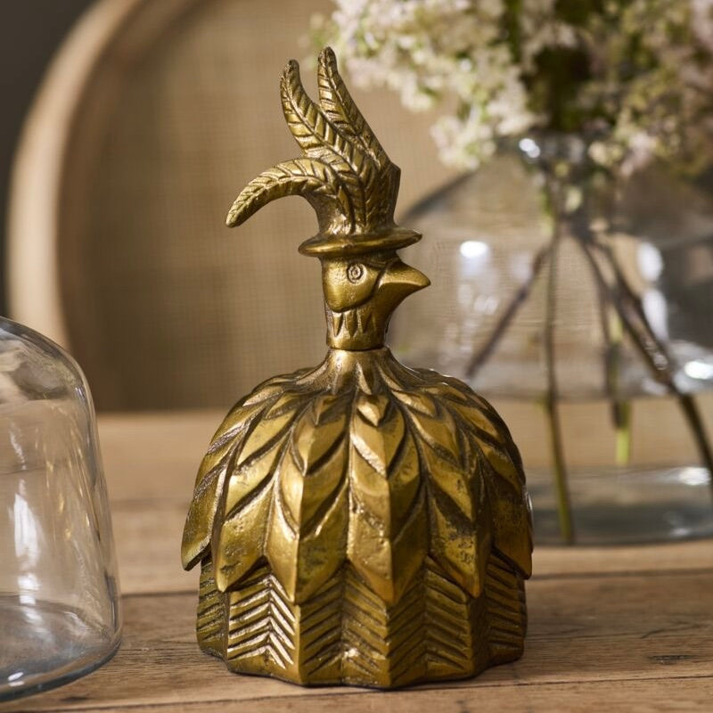A decorative brass rooster figurine with a tall feathered crest and small hat, displayed on a wooden surface with a blurred glass vase and dried flowers in the background.
