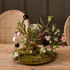 A decorative brass deer head arrangement on a round tray, surrounded by green moss and small wildflowers, set on a wooden table with woven-back chairs in the background.