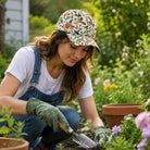 A woman gardening in a sunny garden, wearing a floral-patterned baseball cap, blue denim overalls, a white t-shirt, and green gloves while using a small hand trowel.
