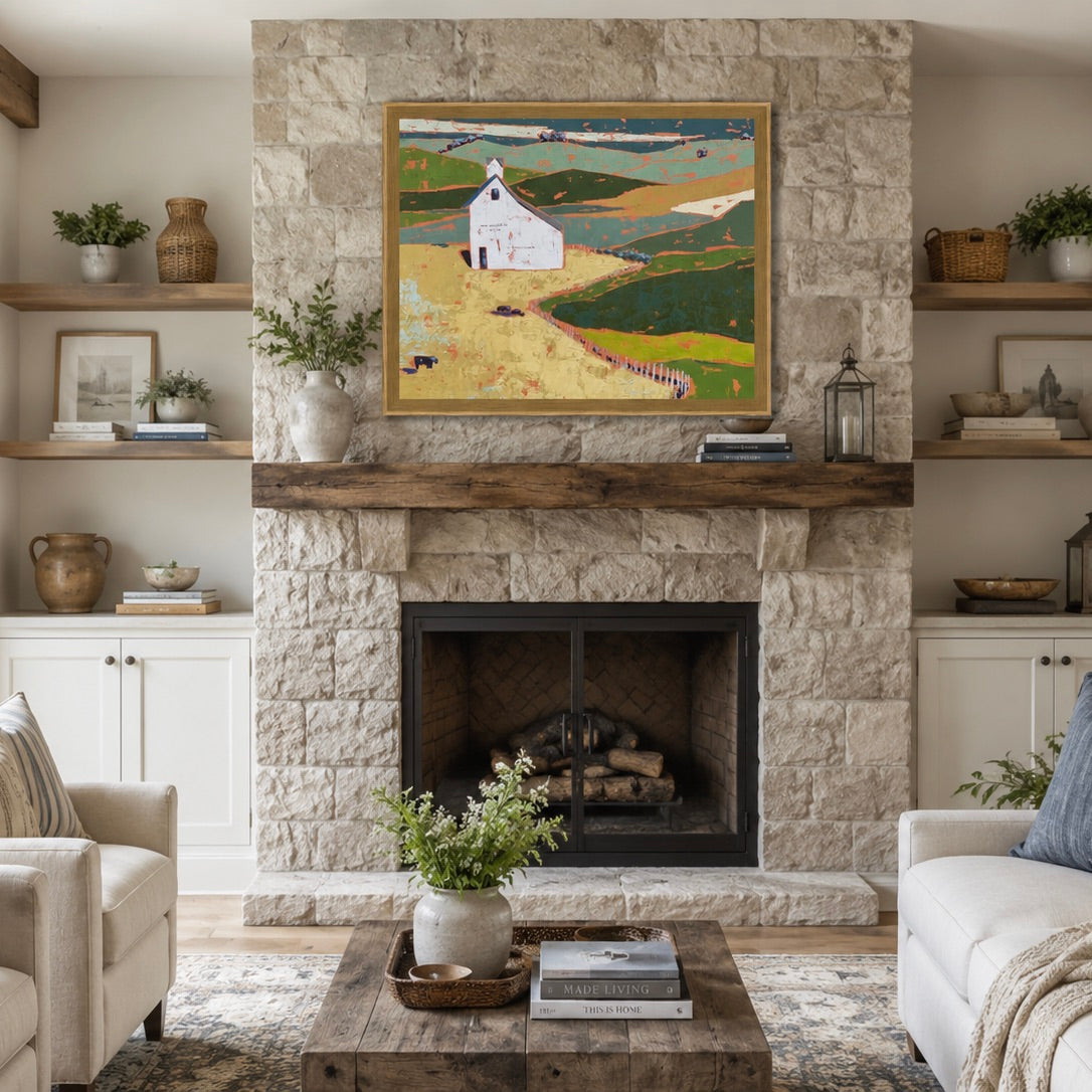 A framed landscape painting of a white church on rolling green hills hangs on a stone fireplace mantel in a styled living room with white walls, built-in shelves, a coffee table, and plants.