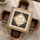 A square framed art print or decorative panel with a central circular design and ornate border details, displayed on a coffee table surrounded by four brown leather chairs in a styled interior setting.