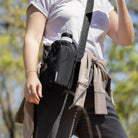 Person wearing a black waist bag with a visible brand logo, standing outdoors with trees in the background.