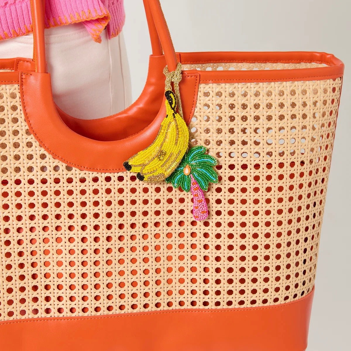 A woven cane tote bag with bright orange leather trim and handles, featuring a colorful embroidered hanging charm shaped like yellow bananas and a green palm tree.