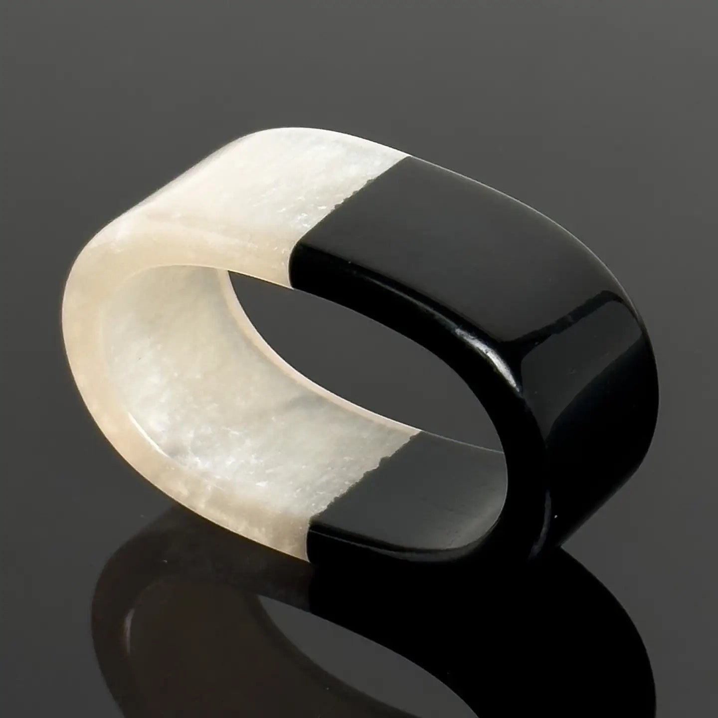 A modern minimalist ring with a split design, featuring a matte off-white stone on one half and a glossy black stone on the other, displayed on a reflective dark surface.