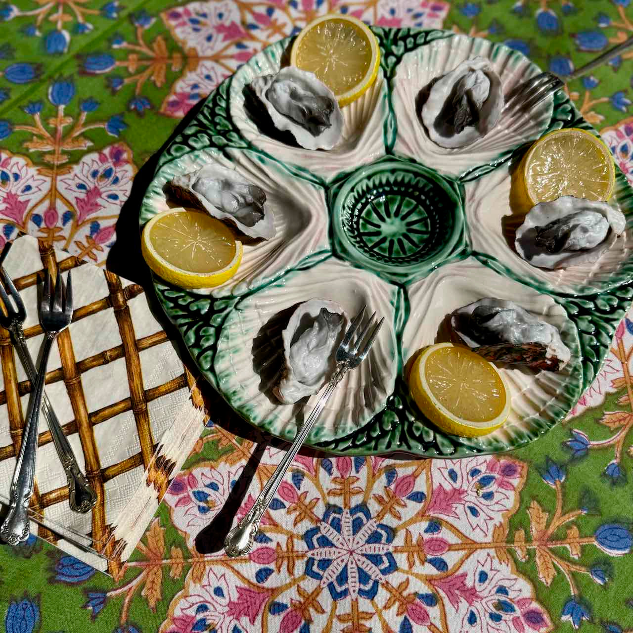 A round green and white ceramic oyster shell platter holding six cooked oysters, each topped with a lemon wedge, set on a colorful patterned tablecloth with forks nearby.