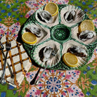 A round green and white ceramic oyster shell platter holding six cooked oysters, each topped with a lemon wedge, set on a colorful patterned tablecloth with forks nearby.