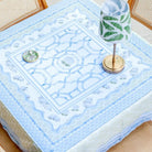 A square tablecloth with a light blue and white geometric pattern draped over a wooden dining table, styled with a green and white patterned table lamp and a small decorative dish.