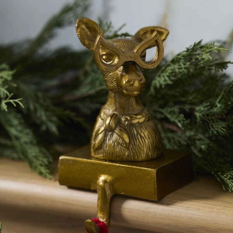 A decorative brass deer figurine with oversized round glasses, mounted on a rectangular base with a hanging hook, styled with green pine branches on a wooden surface.