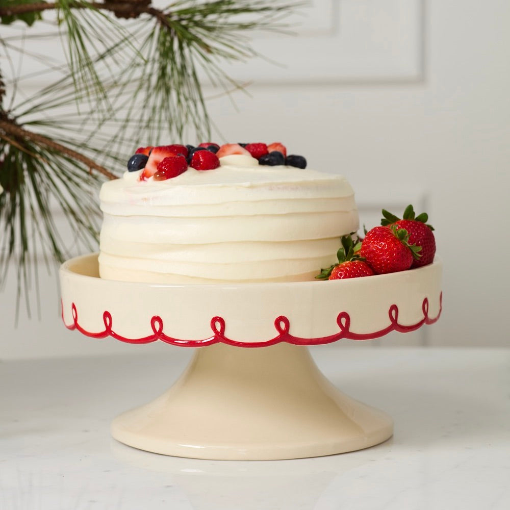 White cake with berries on a decorative cake stand with red patterns, against a neutral background.