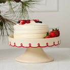White cake with berries on a decorative cake stand with red patterns, against a neutral background.
