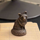 A small metallic bear figurine with a textured, antique bronze finish, sitting on a beige lined notepad with a dark, out-of-focus background.
