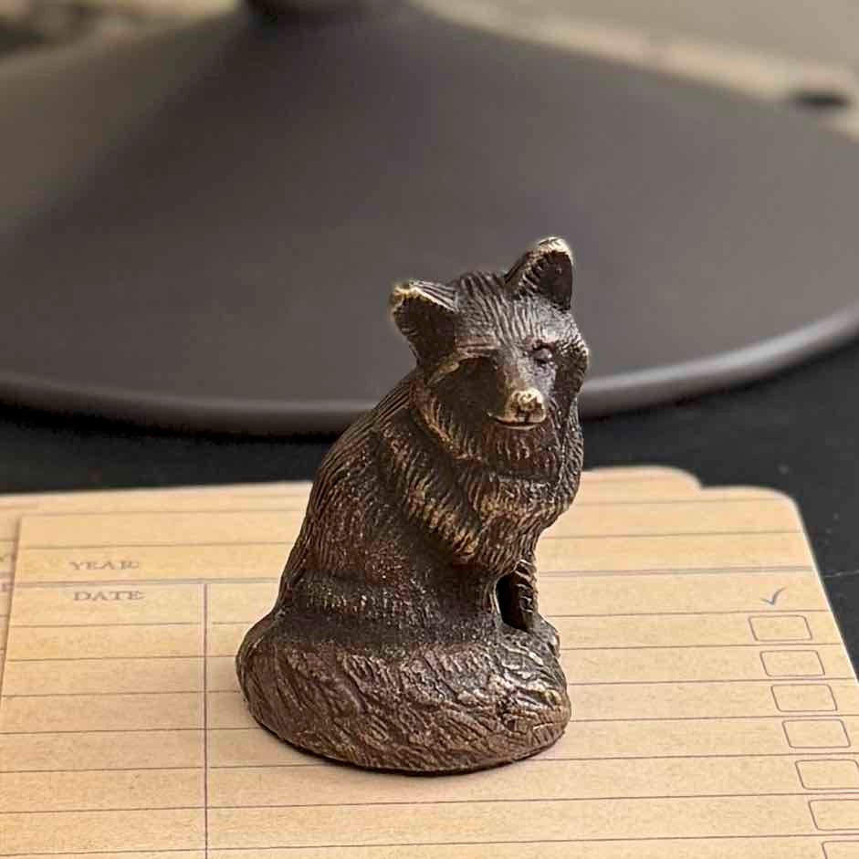 A small metallic bear figurine with a textured, antique bronze finish, sitting on a beige lined notepad with a dark, out-of-focus background.